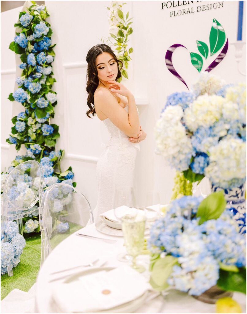 Snohomish Wedding Photographer GSquared Weddings A woman in a white dress poses elegantly beside a floral display of blue hydrangeas. She stands near a table set with blue and white flowers, clear chairs, and elegant tableware. A floral design logo is visible on the wall behind her. Serving Seattle, Snohomish and Orlando