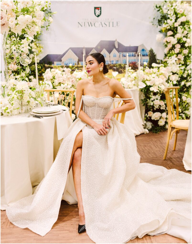 Snohomish Wedding Photographer GSquared Weddings A woman in an elegant strapless white gown with a long train sits gracefully at a decorated table. The background features lush floral arrangements and an image of a grand building. She wears her hair up and has a thoughtful expression. Serving Seattle, Snohomish and Orlando