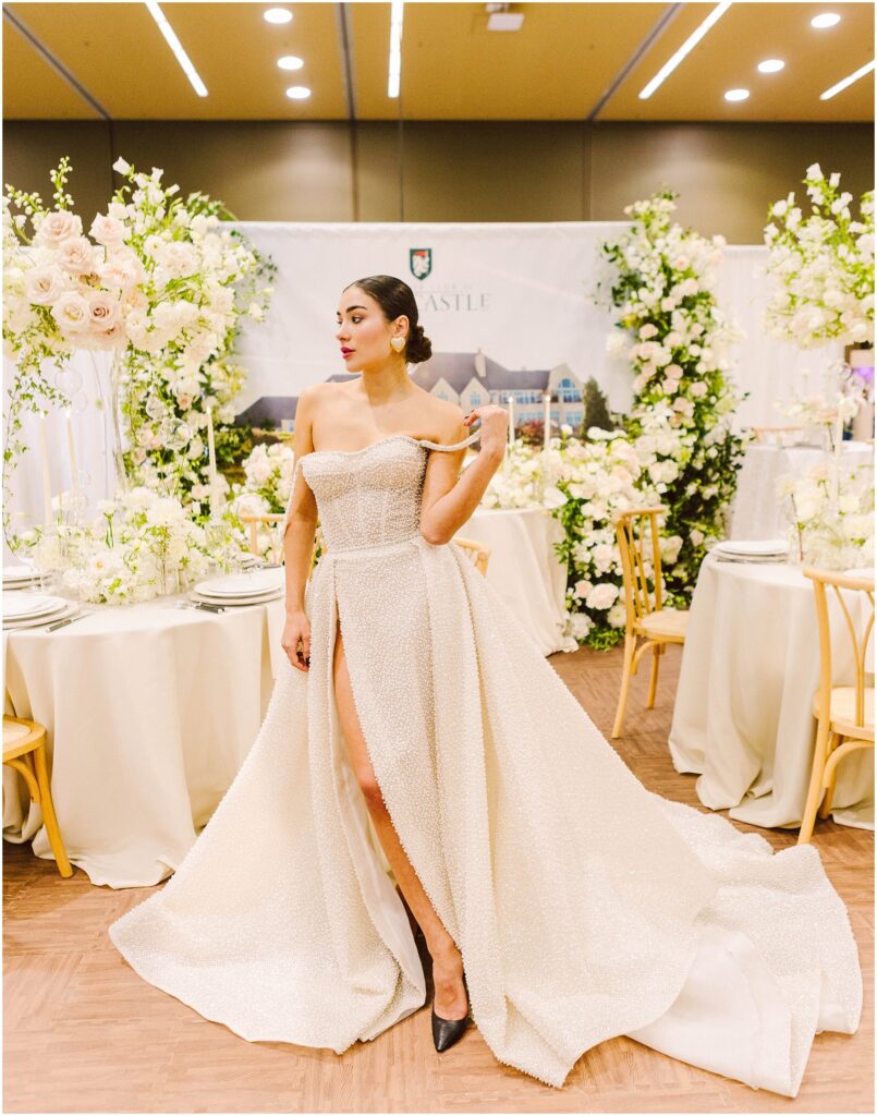 Snohomish Wedding Photographer GSquared Weddings A woman wearing an elegant off-the-shoulder gown with a high slit stands in a floral-decorated venue. She is surrounded by white roses and greenery. The table settings include white linens, with a backdrop featuring architectural imagery. Serving Seattle, Snohomish and Orlando