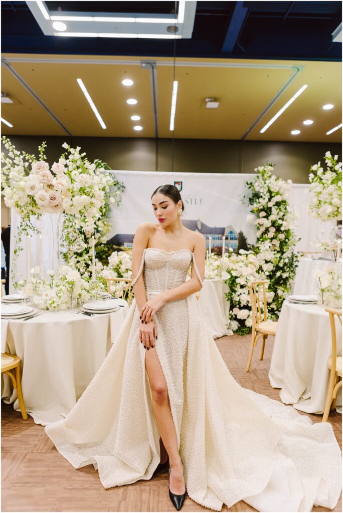 Snohomish Wedding Photographer GSquared Weddings A woman in an elegant white gown with a thigh-high slit poses in a decorated room. She stands in front of tables adorned with white flowers and tall floral arrangements. The setting is luxurious and well-lit. Serving Seattle, Snohomish and Orlando