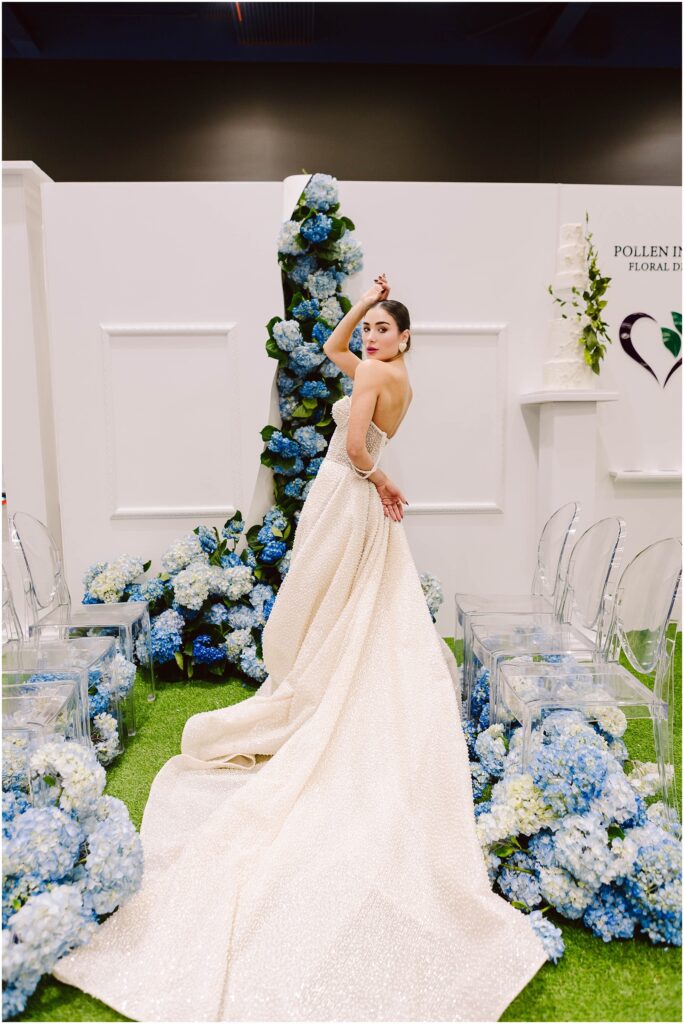 Snohomish Wedding Photographer GSquared Weddings Bride in a sparkling gown poses elegantly among clusters of blue and white hydrangeas. Clear chairs and white walls border the scene, creating a sophisticated wedding setting. Serving Seattle, Snohomish and Orlando