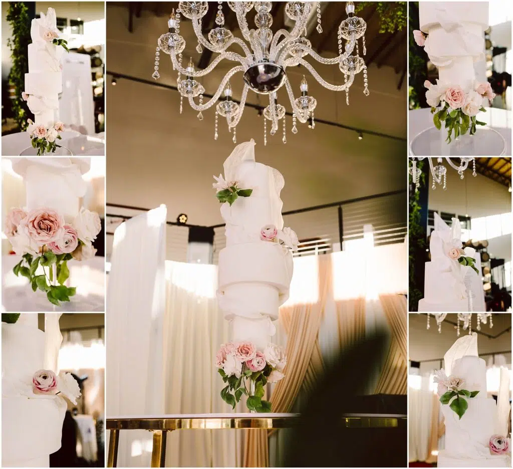 Snohomish Wedding Photographer GSquared Weddings A multi-tiered white wedding cake with elaborate swirling fondant designs, adorned with pink roses. The cake is displayed on a reflective table beneath a sparkling crystal chandelier, surrounded by draped curtains. Serving Seattle, Snohomish and Orlando