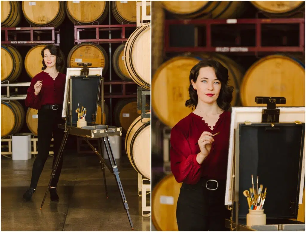 Snohomish Wedding Photographer GSquared Weddings A woman with dark hair, wearing a red blouse and black pants, stands by an easel in a winery or cellar. She holds a brush and poses confidently in front of wooden barrels. Brushes and paints are visible on the easel. Serving Seattle, Snohomish and Orlando