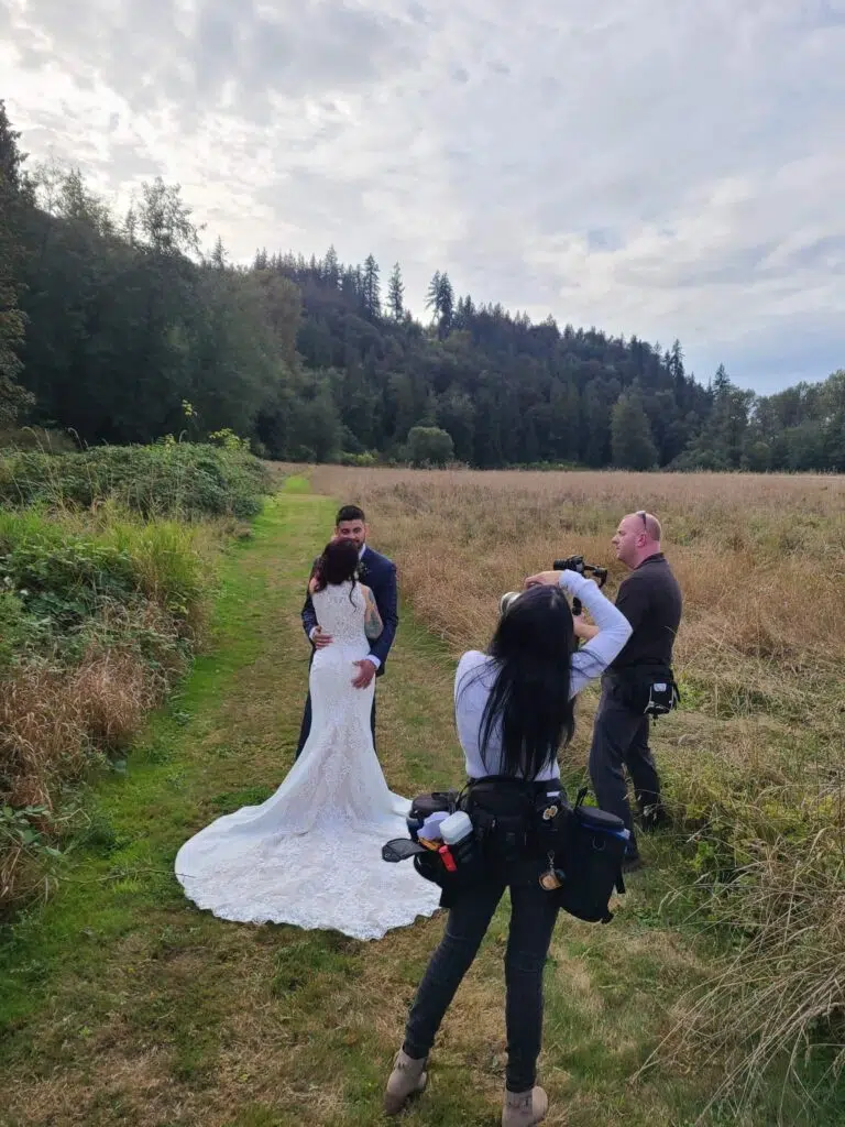 Snohomish Wedding Photographer GSquared Weddings A couple in wedding attire embraces on a grassy path surrounded by tall grass and trees. A photographer and assistant capture the moment. The sky is cloudy, and a forested hillside is in the background. Serving Seattle, Snohomish and Orlando