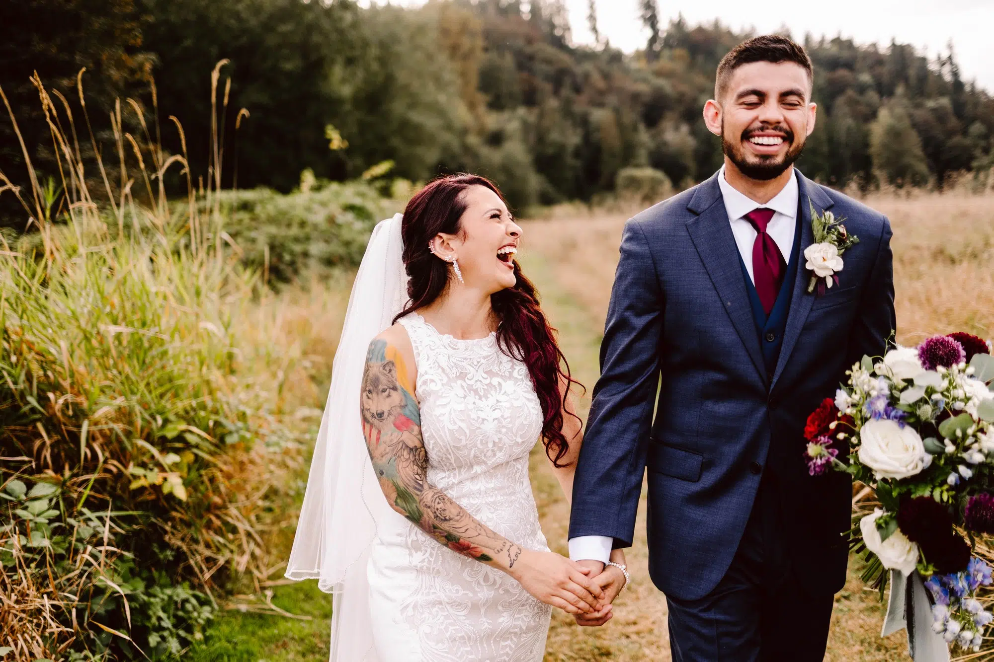 Snohomish Wedding Photographer GSquared Weddings A bride and groom joyfully walk handinhand through a grassy field effortlessly posing for their special moment The bride in a lace dress and veil flaunts colorful tattoos as she laughs at the groom in his blue suit holding a bouquet Trees and bushes frame this blissful scene Serving Seattle Snohomish and Orlando Snohomish Wedding Photographer GSquared Weddings A bride and groom joyfully walk hand in hand through a grassy field effortlessly posing for their special moment The bride in a lace dress and veil flaunts colorful tattoos as she laughs at the groom in his blue suit holding a bouquet Trees and bushes frame this blissful scene Serving Seattle Snohomish and Orlando