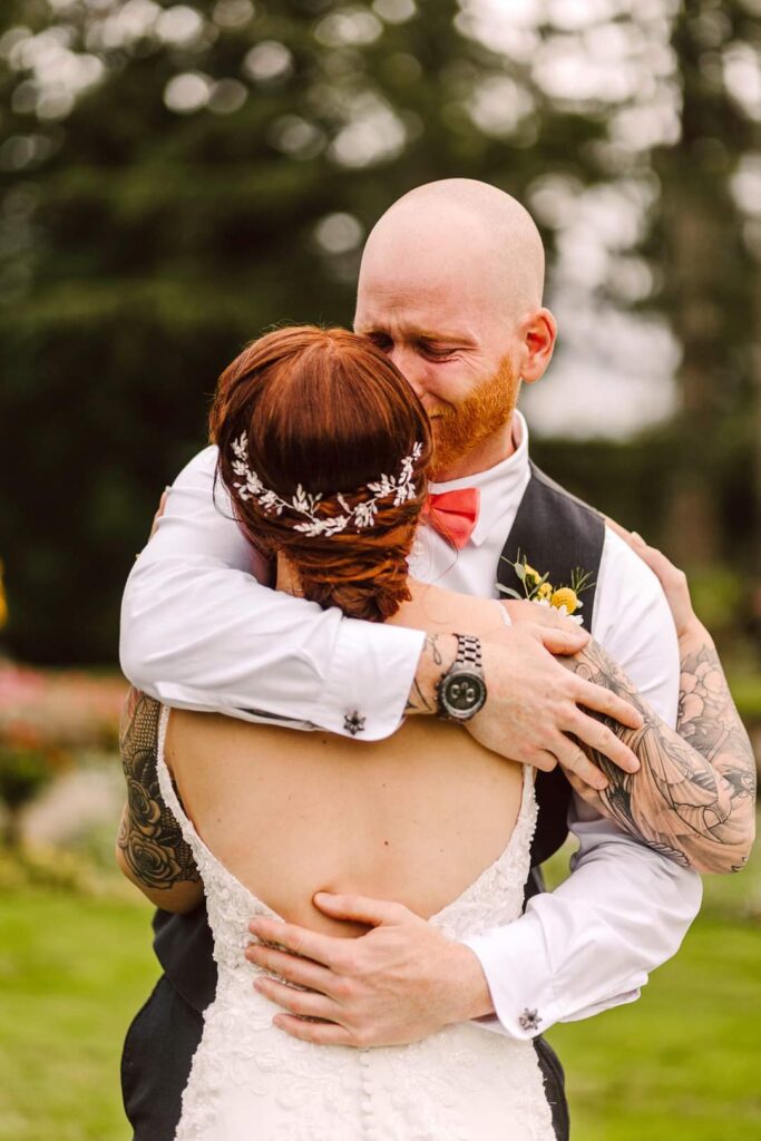 Snohomish Wedding Photographer GSquared Weddings A bald man in a vest tenderly embraces a woman in a white dress adorned with an intricate hair accessory. Outdoors, amidst lush greenery, he appears on the verge of tears, his eyes closed and face showing a deeply emotional expression. Serving Seattle, Snohomish and Orlando