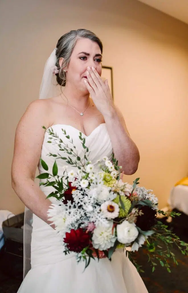 Snohomish Wedding Photographer GSquared Weddings A bride in a white dress and veil stands indoors, holding a bouquet of white, red, and green flowers. She appears emotional, on the verge of tears as she covers her mouth with one hand. Serving Seattle, Snohomish and Orlando