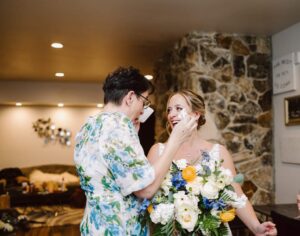 Snohomish Wedding Photographer GSquared Weddings Two women joyfully converse in a warmly lit room. One wears glasses and a floral dress, while the other, in a white dress holding a bouquet of flowers, seems to be celebrating her wedding. The stone wall behind them adds to the charm of this special occasion. Serving Seattle, Snohomish and Orlando