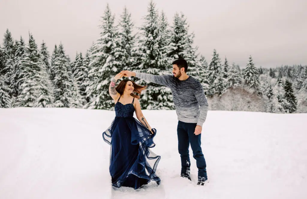 Snohomish Wedding Photographer GSquared Weddings A couple twirls joyfully in a snowy landscape, immortalized in stunning photos. The woman wears a flowing navy blue gown, while the man dons a gray sweater and dark pants. Snow-covered trees and a cloudy sky complete the enchanting backdrop. Serving Seattle, Snohomish and Orlando