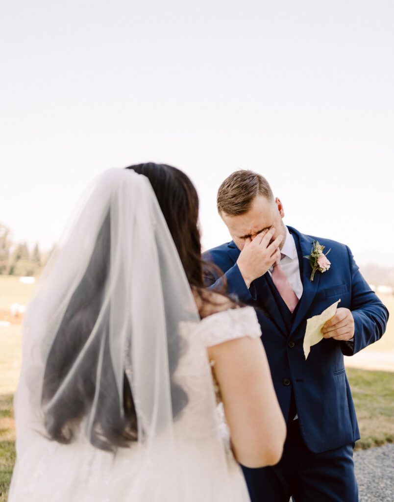 Snohomish Wedding Photographer GSquared Weddings A groom in a blue suit wipes his teary eyes while holding a handwritten note, standing outdoors. A bride, with long hair and a veil, faces him in her white dress. The background reveals a grassy field under a clear sky. Serving Seattle, Snohomish and Orlando