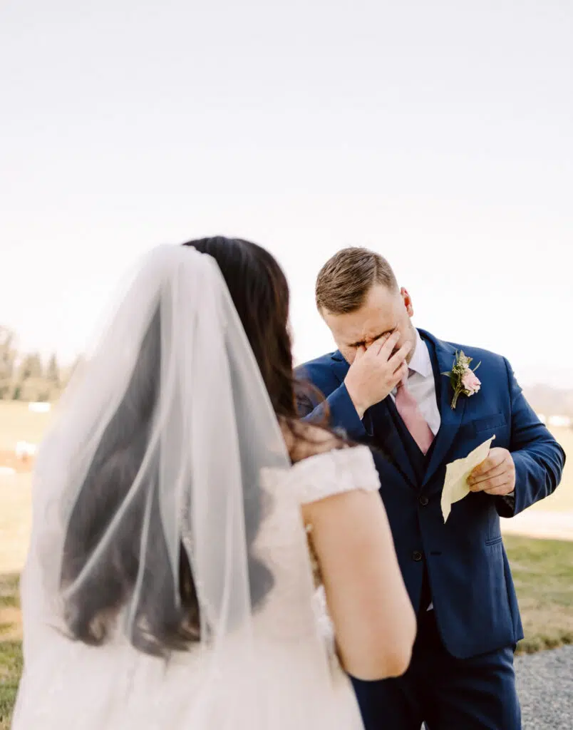 Snohomish Wedding Photographer GSquared Weddings A groom in a blue suit wipes his teary eyes while holding a handwritten note, standing outdoors. A bride, with long hair and a veil, faces him in her white dress. The background reveals a grassy field under a clear sky. Serving Seattle, Snohomish and Orlando