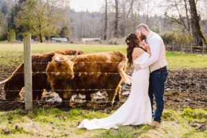 Bride and groom kissing near fence with cattle and rural landscape in the background.