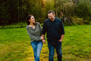 Snohomish Wedding Photographer GSquared Weddings A couple is walking hand in hand on a grassy field surrounded by trees. The woman, wearing a green sweater, smiles and looks at the man, who is wearing a dark blue shirt. Sunlight filters through the trees in the background. Serving Seattle, Snohomish and Orlando