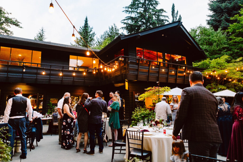 Snohomish Wedding Photographer GSquared Weddings A lively outdoor gathering at a house features guests mingling near tables with lights strung above. The backdrop is a large two-story building surrounded by trees. Everyone is dressed in formal attire, suggesting a celebratory event or party. Serving Seattle, Snohomish and Orlando