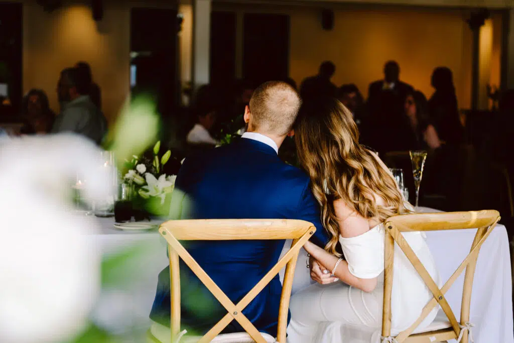 Snohomish Wedding Photographer GSquared Weddings A couple sits closely together on wooden chairs at a wedding reception. The man wears a blue suit, and the woman dazzles in an off-shoulder white dress. Theyre holding hands, facing away from the camera, with blurred guests and elegant decor in the background. Serving Seattle, Snohomish and Orlando