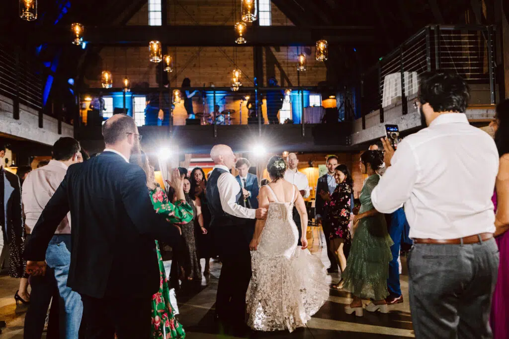 Snohomish Wedding Photographer GSquared Weddings A newlywed couple dances in the center of a rustic, warmly lit reception hall, surrounded by delighted guests. Some clap along while others capture photos, all beneath high ceilings adorned with hanging lights and an inviting open upper level. Serving Seattle, Snohomish and Orlando