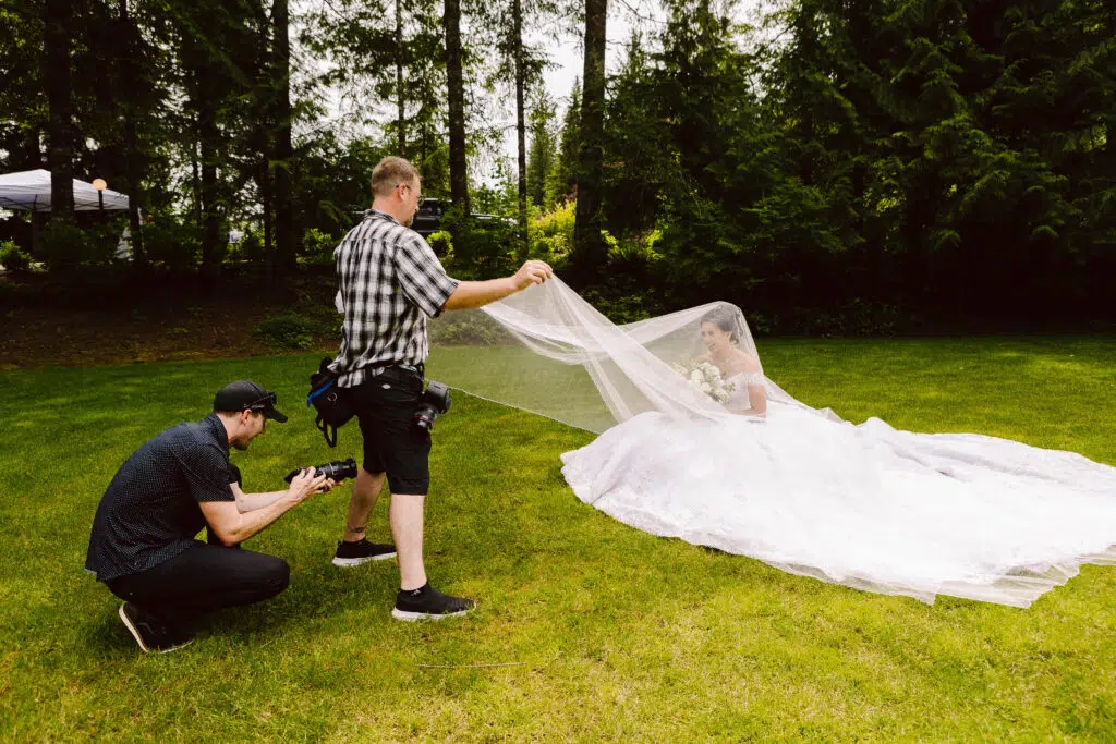 Snohomish Wedding Photographer GSquared Weddings A talented photographer kneels to capture a stunning image of a bride in her white dress sitting gracefully on the grass. Meanwhile, an assistant meticulously adjusts her veil. They find themselves enveloped by natures beauty, with tall trees and lush greenery all around. Serving Seattle, Snohomish and Orlando