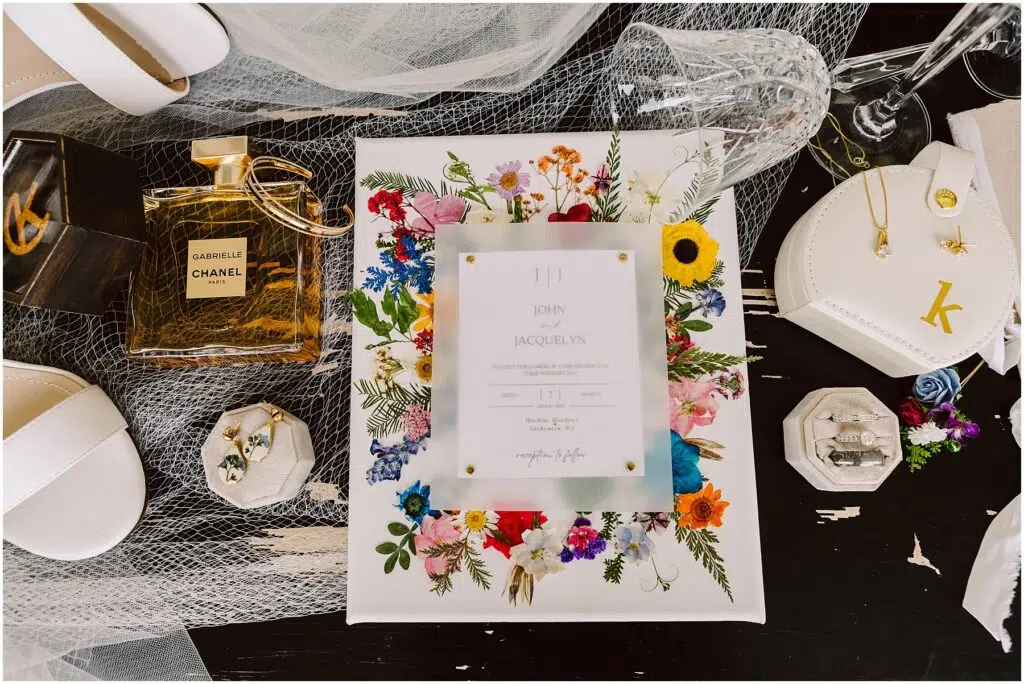 Snohomish Wedding Photographer GSquared Weddings A flat lay image showcases wedding invitations amidst a vibrant floral design, alongside white shoes, Chanel perfume, jewelry, and rings in a box on dark machias. Nearby rests a heart-shaped case with a K, all beautifully arranged on tulle. Serving Seattle, Snohomish and Orlando