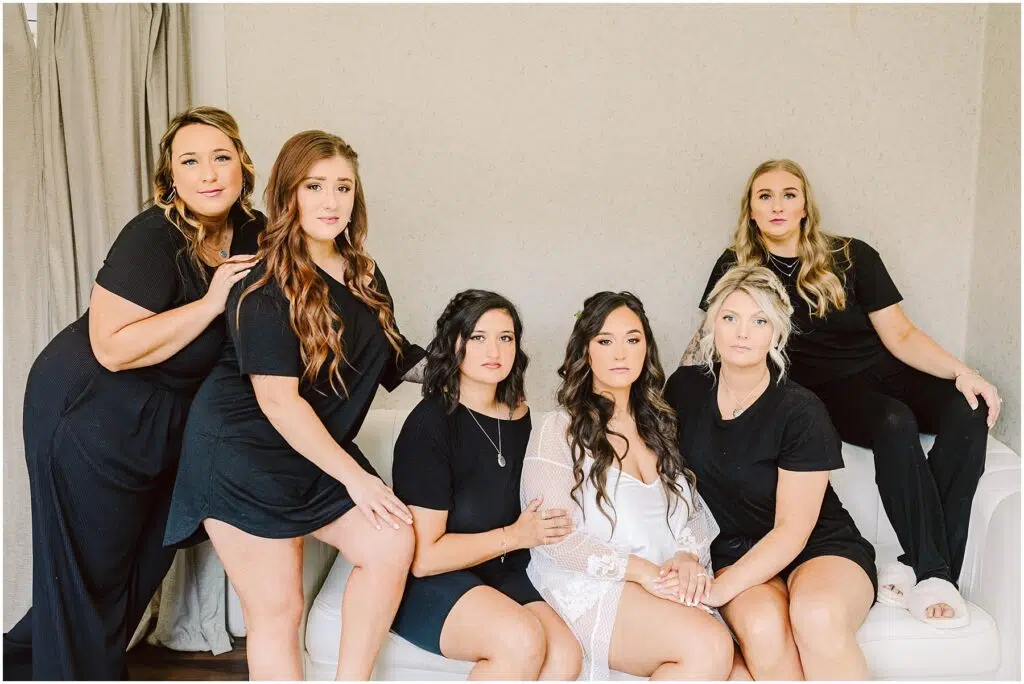 Snohomish Wedding Photographer GSquared Weddings A group of six women poses indoors in Machias. The woman in the center dons a sheer white robe, while the others wear black outfits. Arranged around a white sofa, they look at the camera with serious expressions, capturing a striking contrast and an aura of elegance. Serving Seattle, Snohomish and Orlando