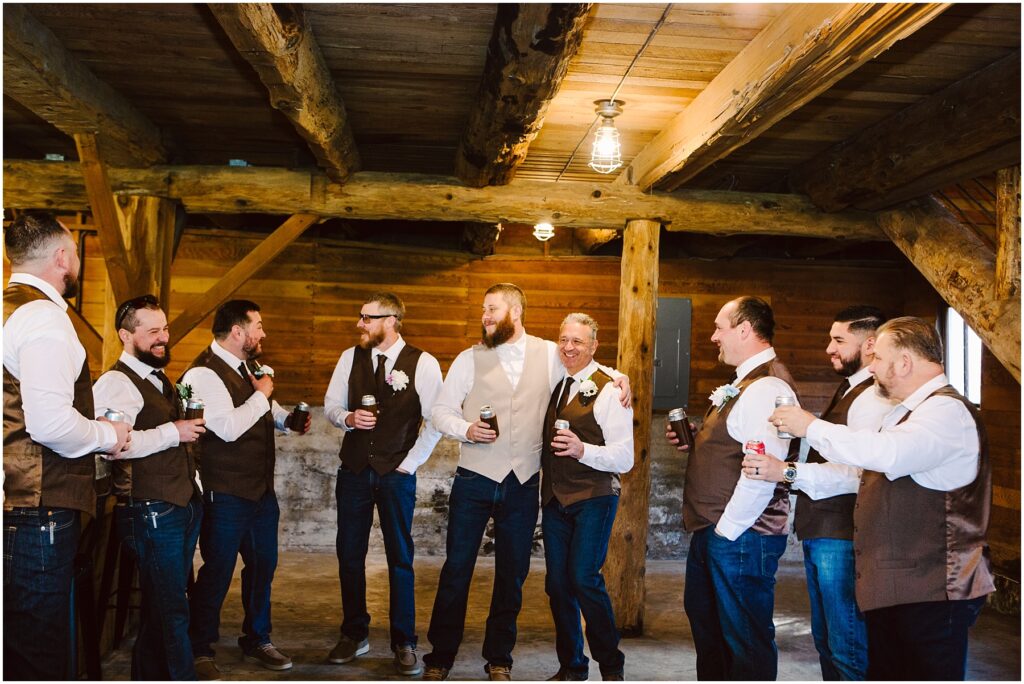 Snohomish Wedding Photographer GSquared Weddings A group of eight men in vests and jeans are standing in a rustic wooden room in Machias, smiling and holding drink bottles. They appear to be at a casual gathering or celebration, possibly a wedding. The mood is relaxed and joyful. Serving Seattle, Snohomish and Orlando