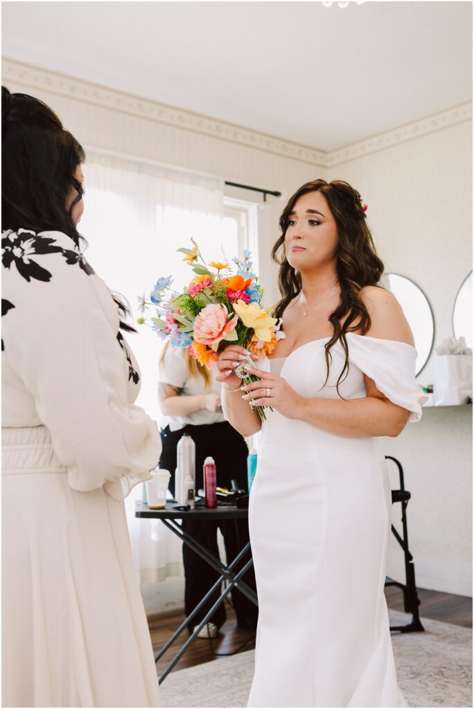 Snohomish Wedding Photographer GSquared Weddings In a charming Machias setting, a bride in an off-shoulder white dress holds a colorful bouquet, facing someone in a cream outfit. Indoors, with salon tools visible behind them, natural light streams through sheer curtains for an ethereal touch. Serving Seattle, Snohomish and Orlando