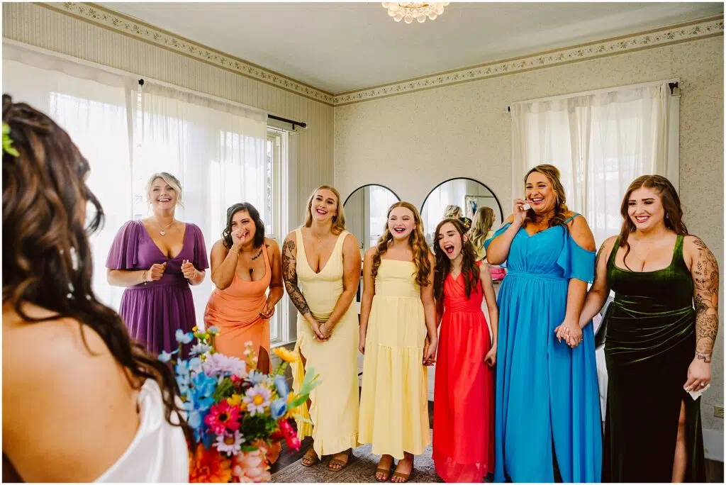 Snohomish Wedding Photographer GSquared Weddings A bride stands with her back to the camera holding a colorful bouquet. In front of her, a group of six bridesmaids and a flower girl from Machias react with joy and surprise. They are wearing dresses in various colors, and the room is decorated with mirrors and curtains. Serving Seattle, Snohomish and Orlando