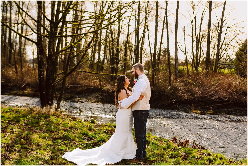 Snohomish Wedding Photographer GSquared Weddings A couple in wedding attire embraces by the scenic Machias River, surrounded by bare trees and greenery. The sun filters through the branches, creating a peaceful and romantic atmosphere. Serving Seattle, Snohomish and Orlando