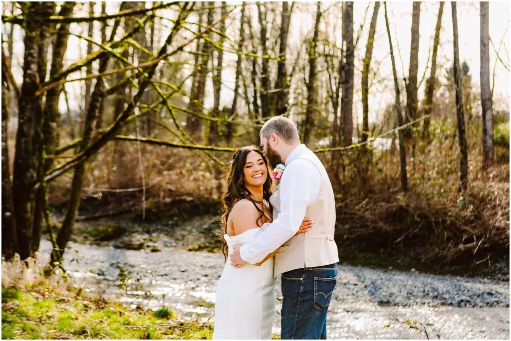 Snohomish Wedding Photographer GSquared Weddings A couple embraces near a small stream in a wooded area reminiscent of Machias. The woman, smiling in her white dress, is held gently by the man in a light shirt and jeans. Leafless trees frame their moment on this sunny day. Serving Seattle, Snohomish and Orlando