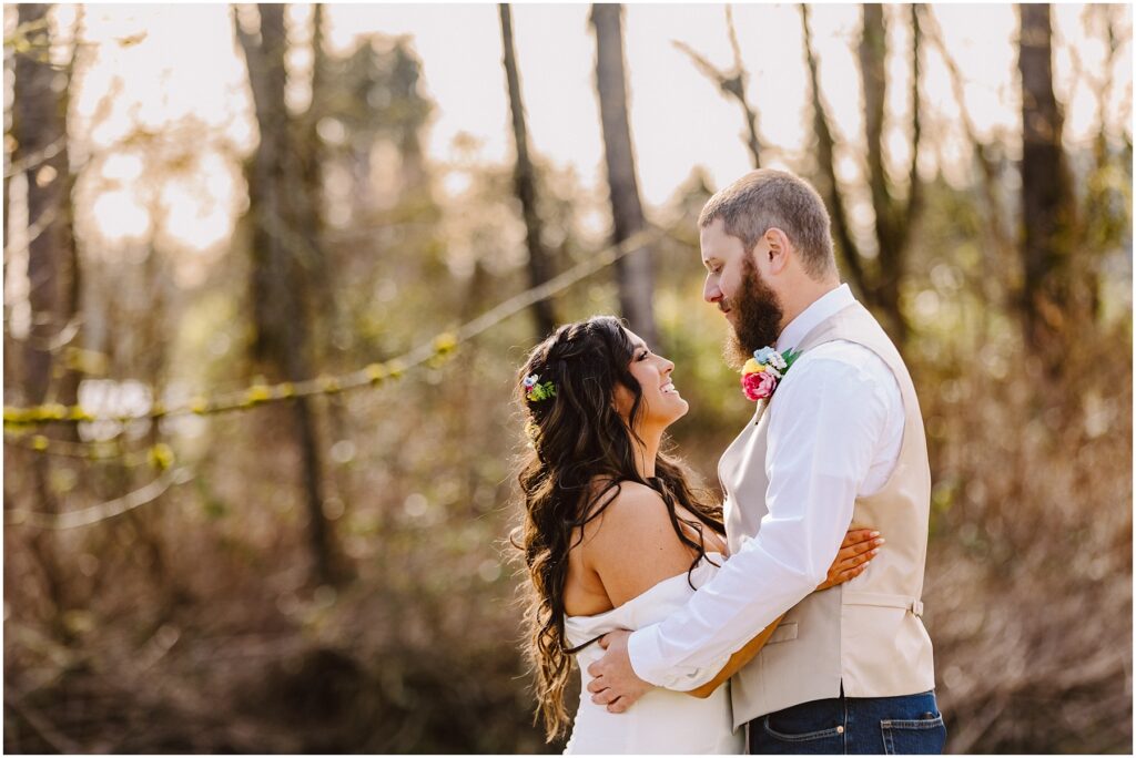Snohomish Wedding Photographer GSquared Weddings In a serene Machias forest clearing, a couple embraces. The woman, adorned with long curly hair and a floral headpiece, wears a white dress. Beside her stands the man, bearded and donning a white shirt and vest with a colorful boutonniere. Sunlight filters through the trees behind them. Serving Seattle, Snohomish and Orlando