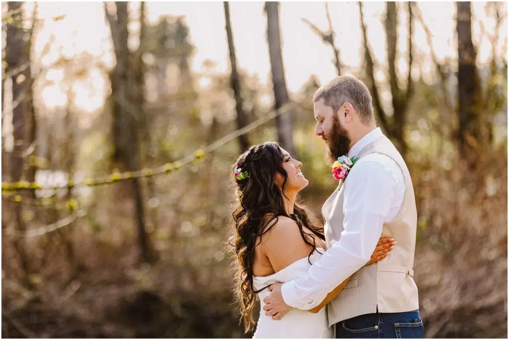 Snohomish Wedding Photographer GSquared Weddings In a serene Machias forest clearing, a couple embraces. The woman, adorned with long curly hair and a floral headpiece, wears a white dress. Beside her stands the man, bearded and donning a white shirt and vest with a colorful boutonniere. Sunlight filters through the trees behind them. Serving Seattle, Snohomish and Orlando