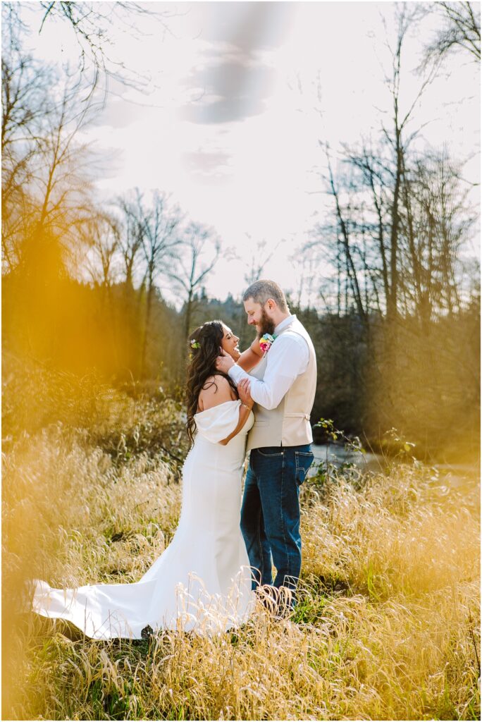 Snohomish Wedding Photographer GSquared Weddings A bride in a white dress and a groom in casual attire stand lovingly in a sunlit, grassy field near Machias. They are surrounded by tall trees and framed by soft, blurred foliage. The groom adjusts the brides hair, creating an intimate and tender moment. Serving Seattle, Snohomish and Orlando