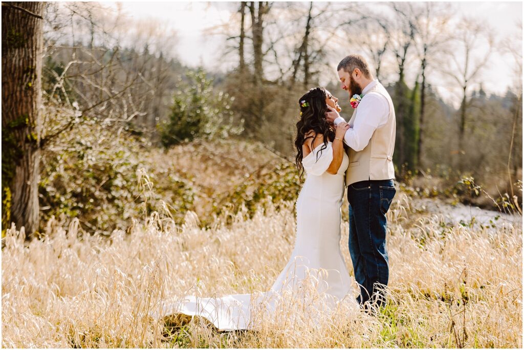 Snohomish Wedding Photographer GSquared Weddings In a field near Machias, the bride in a white off-the-shoulder dress and the groom in a vest and jeans stand amidst tall grasses. With trees as their backdrop, they face each other closely, creating an enchanting and romantic scene. Serving Seattle, Snohomish and Orlando