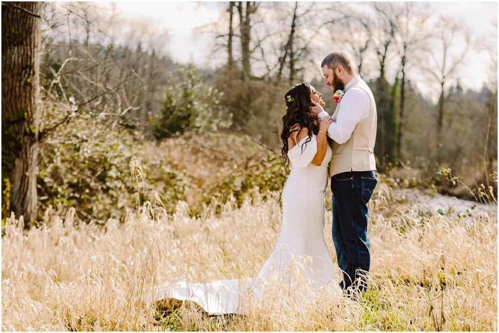 Snohomish Wedding Photographer GSquared Weddings In a field near Machias, the bride in a white off-the-shoulder dress and the groom in a vest and jeans stand amidst tall grasses. With trees as their backdrop, they face each other closely, creating an enchanting and romantic scene. Serving Seattle, Snohomish and Orlando