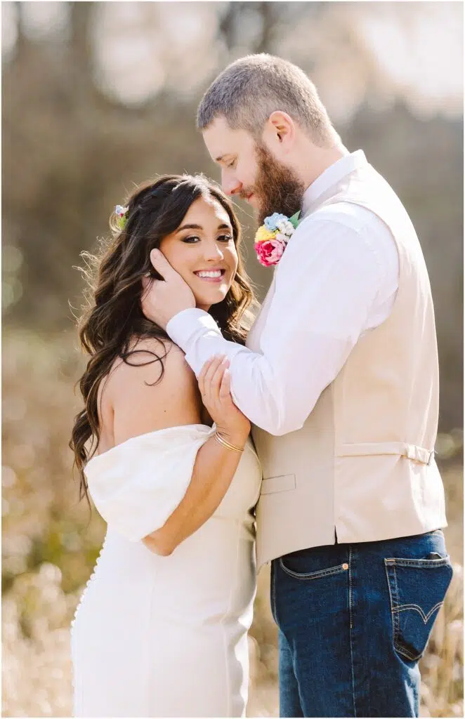 Snohomish Wedding Photographer GSquared Weddings A couple stands in Machias, embracing happily. The woman smiles at the camera in a white dress with floral accents in her hair. The bearded man gently touches her face, wearing a light vest over a white shirt. Sunlight filters through the background, capturing their joy. Serving Seattle, Snohomish and Orlando