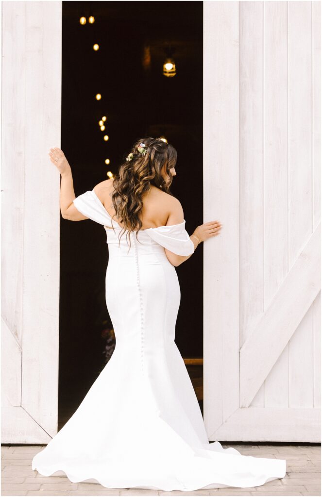 Snohomish Wedding Photographer GSquared Weddings A bride in an off-shoulder white gown stands facing a large, open barn door decorated with string lights. Her long hair is styled with flowers, and she looks back slightly in the serene Machias countryside, highlighting the gowns button details and flowing train. Serving Seattle, Snohomish and Orlando
