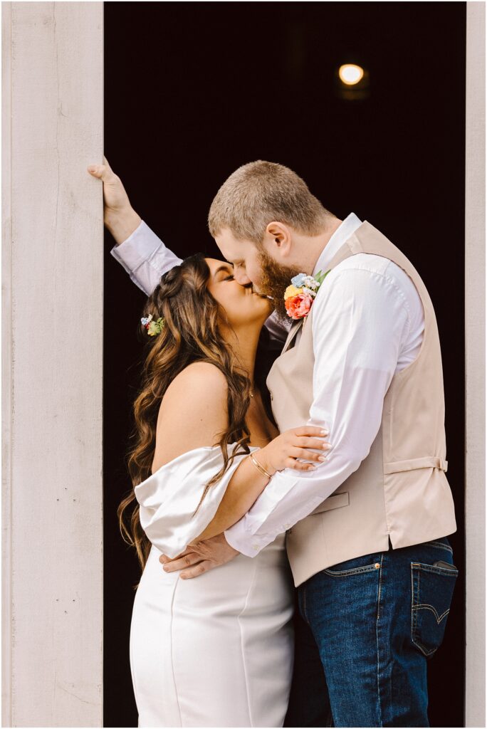 Snohomish Wedding Photographer GSquared Weddings In the charming town of Machias, a bride in an off-shoulder white dress and a groom in a beige vest and jeans share a kiss. They stand in a doorway, with floral details adorning them and flowers nestled in her hair, as his arm rests against the frame. Serving Seattle, Snohomish and Orlando
