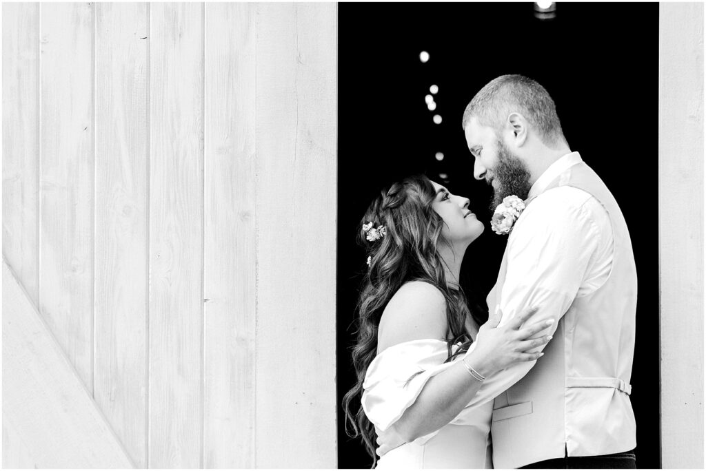Snohomish Wedding Photographer GSquared Weddings A couple embraces lovingly in a Machias doorway. The woman looks up at the man, smiling. Both are dressed in wedding attire, with the man wearing a vest and the woman having flowers in her hair. The scene is beautifully captured in black and white. Serving Seattle, Snohomish and Orlando