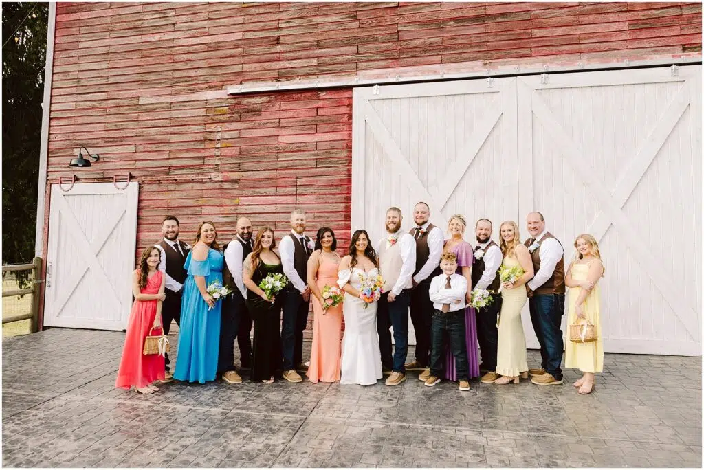 Snohomish Wedding Photographer GSquared Weddings A wedding party gathers joyfully in front of a charming red barn with large white doors, reminiscent of quaint Machias architecture. The group, dressed in formal attire, includes men in vests and women in colorful dresses. A young boy and girl are also part of the cheerful group, all smiling radiantly. Serving Seattle, Snohomish and Orlando
