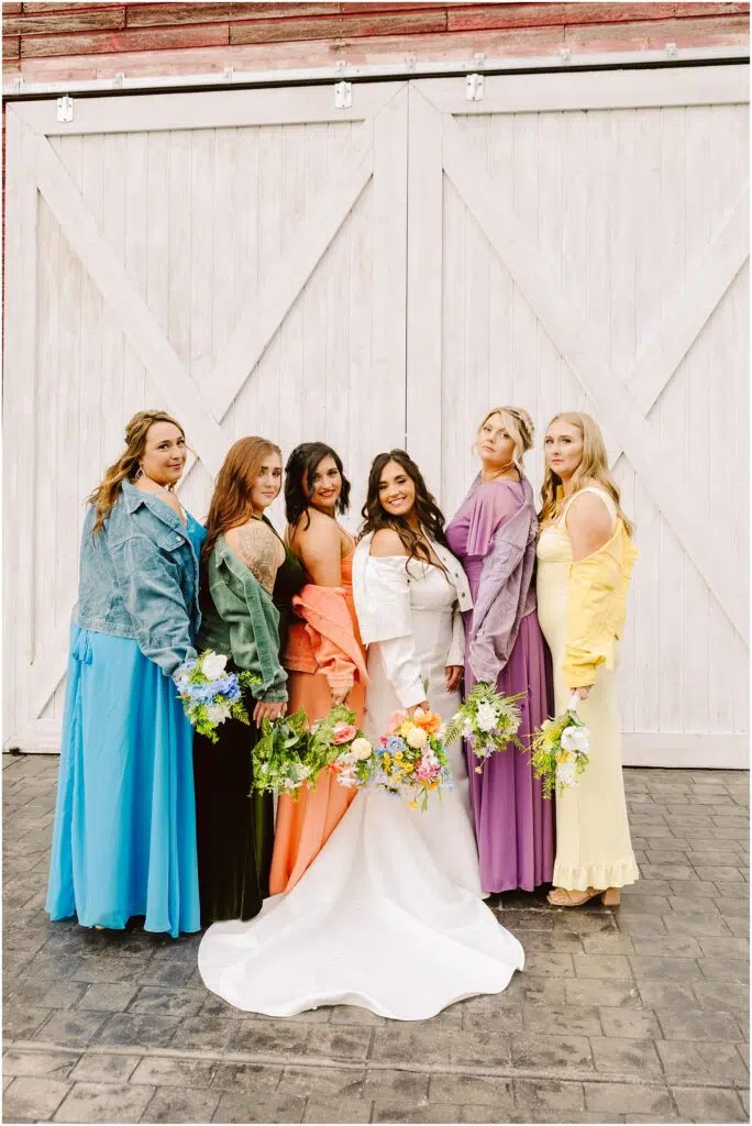 Snohomish Wedding Photographer GSquared Weddings A bride in a white dress stands with five bridesmaids in colorful dresses and denim jackets, all gathered in front of Machiass iconic large white wooden doors. Each holds a bouquet of flowers, arranged in a row facing the camera, smiling. Serving Seattle, Snohomish and Orlando