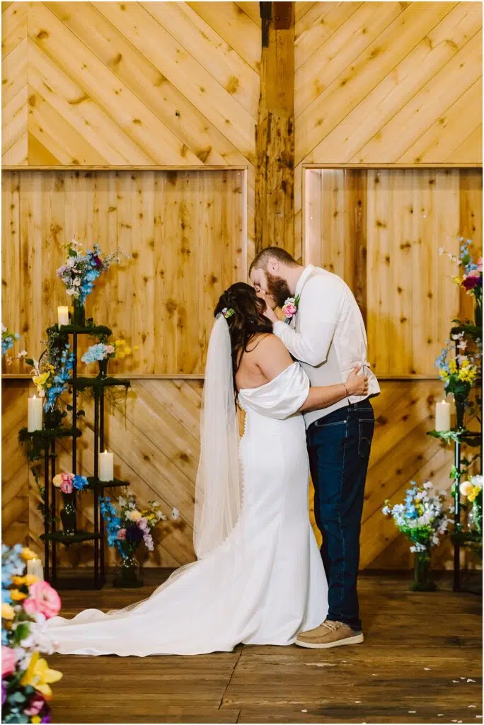 Snohomish Wedding Photographer GSquared Weddings In a rustic Machias setting, a couple shares a kiss among wooden walls and colorful floral arrangements. The bride wears a white gown and veil, while the groom sports a white shirt and jeans. Candles and vibrant flowers enhance the romantic atmosphere. Serving Seattle, Snohomish and Orlando