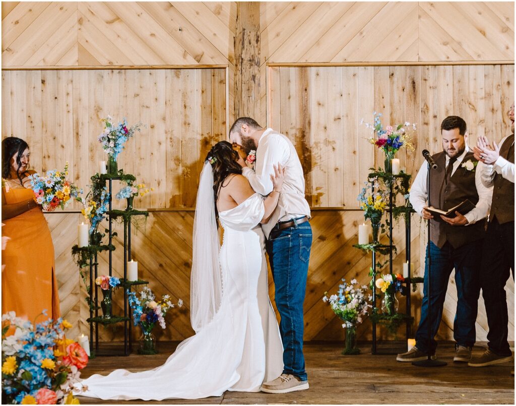 Snohomish Wedding Photographer GSquared Weddings A bride and groom share a kiss during their Machias wedding ceremony in front of a wooden backdrop. The groom is wearing jeans and a white shirt; the bride is in an off-shoulder white dress. They are surrounded by colorful flowers and guests clapping. Serving Seattle, Snohomish and Orlando