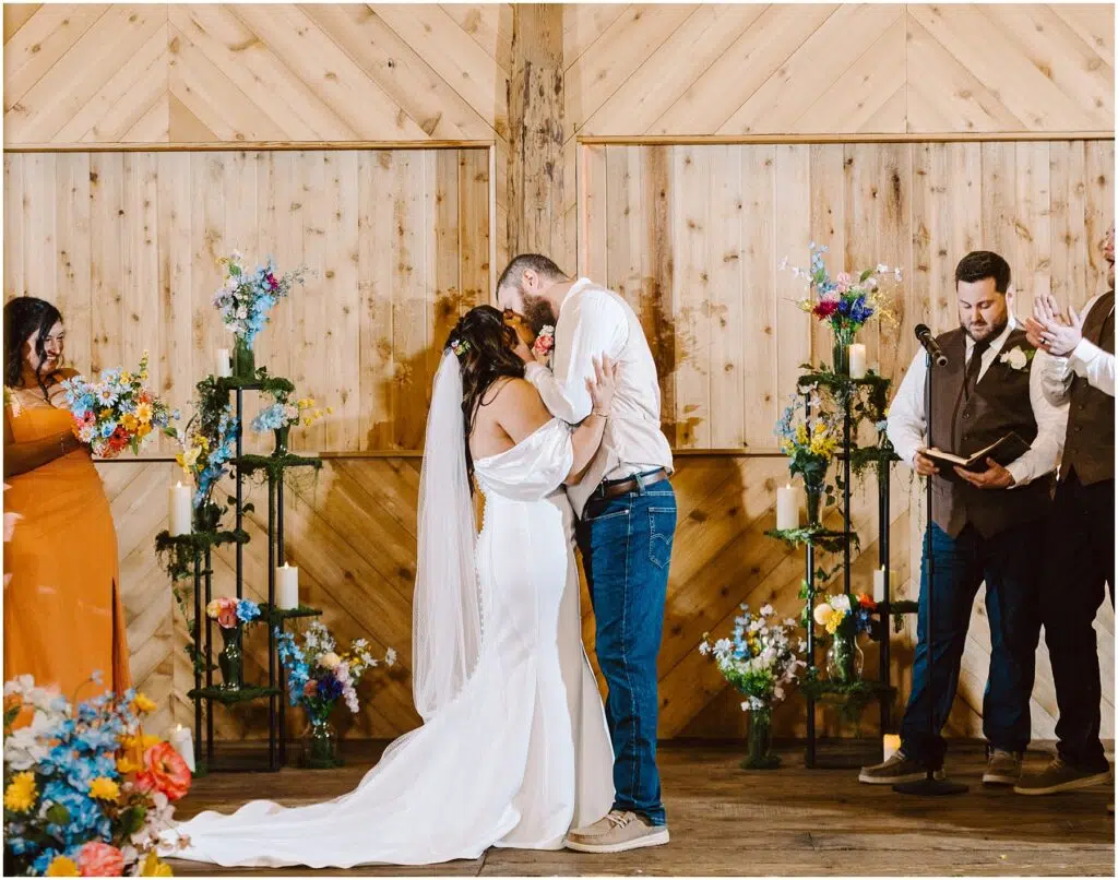 Snohomish Wedding Photographer GSquared Weddings A bride and groom share a kiss during their Machias wedding ceremony in front of a wooden backdrop. The groom is wearing jeans and a white shirt; the bride is in an off-shoulder white dress. They are surrounded by colorful flowers and guests clapping. Serving Seattle, Snohomish and Orlando