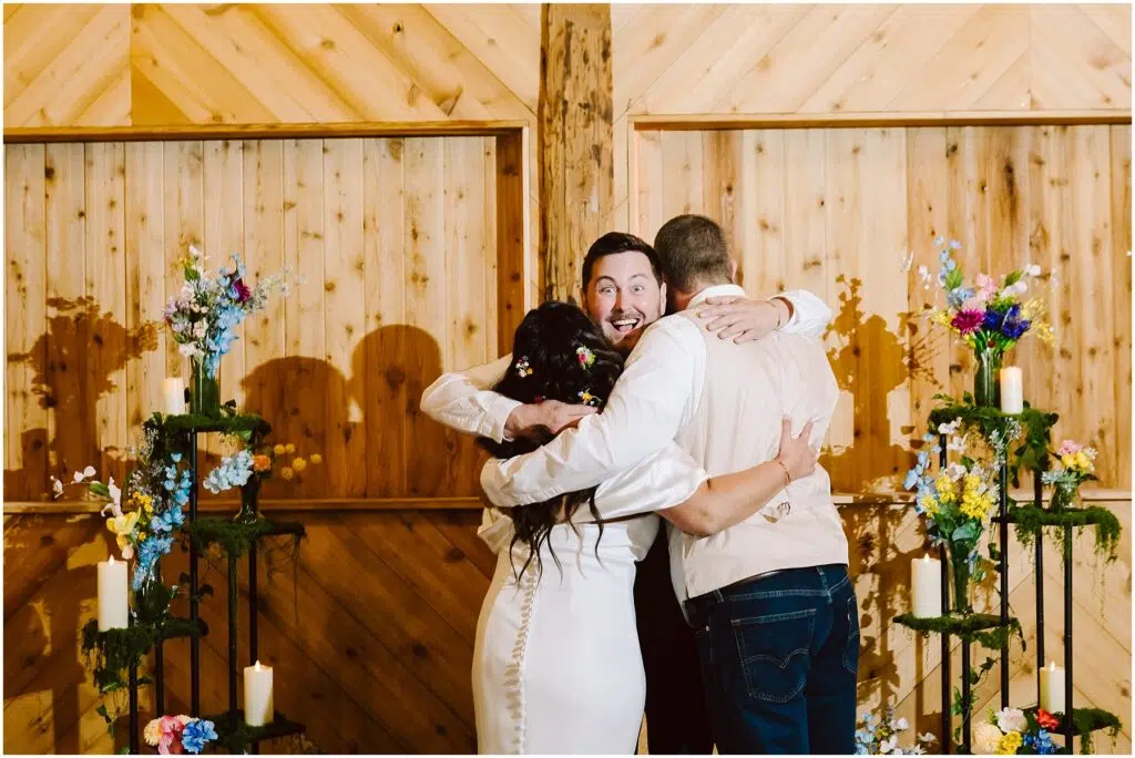 Snohomish Wedding Photographer GSquared Weddings Three people in a group hug at a wedding, standing in front of a wooden Machias wall. Flowers and candles decorate the surroundings, casting a warm glow. The person in the middle, wearing a vest, has a joyful expression. Serving Seattle, Snohomish and Orlando