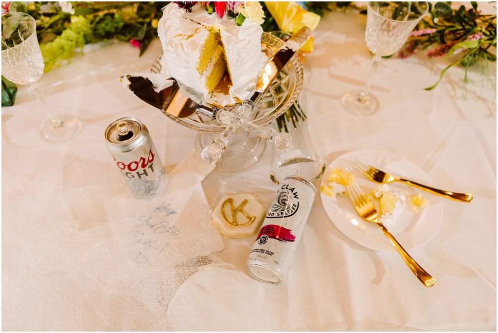 Snohomish Wedding Photographer GSquared Weddings A table in Machias showcases a sliced white frosted cake on a glass stand, flanked by a Coors Light can, a White Claw can, champagne flutes, a plate with cake, and gold utensils. Decorative flowers and greenery provide an elegant backdrop. Serving Seattle, Snohomish and Orlando
