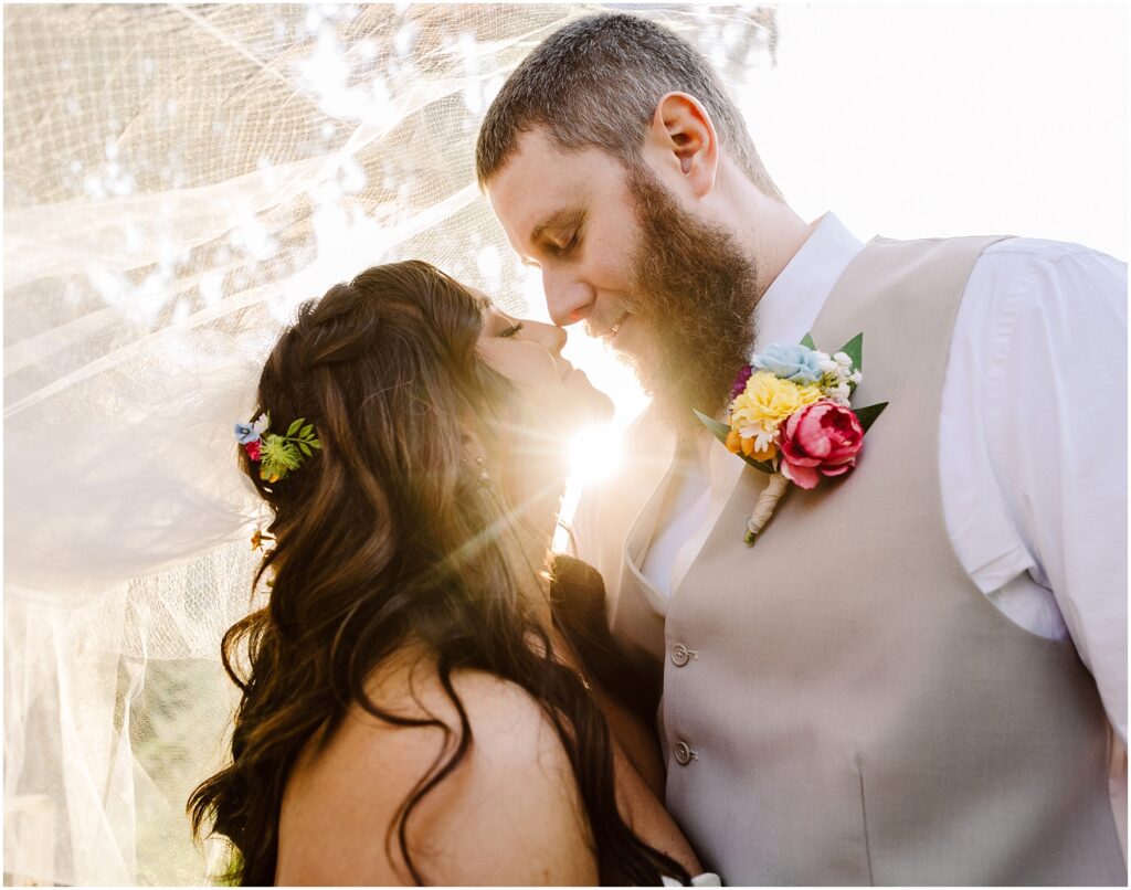 Snohomish Wedding Photographer GSquared Weddings Bride and groom embrace under a veil with the sun shining behind them, creating a moment as enchanting as a Machias sunset. The grooms beige vest complements his floral boutonniere, while the brides hair blooms with flowers. They gaze at each other lovingly. Serving Seattle, Snohomish and Orlando