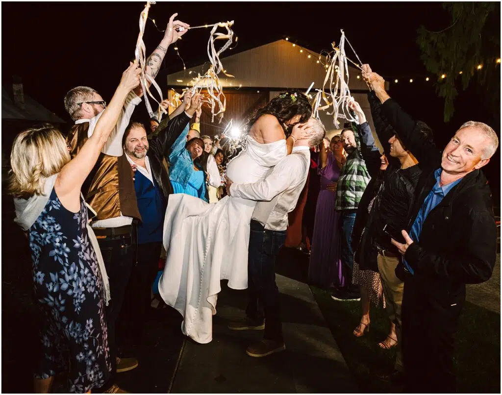 Snohomish Wedding Photographer GSquared Weddings Amid the joyful crowd in Machias, friends wave ribbon wands under the night sky. The man lifts the woman triumphantly as guests form a festive arch with ribbons, their warm laughter blending seamlessly with the celebration. Serving Seattle, Snohomish and Orlando