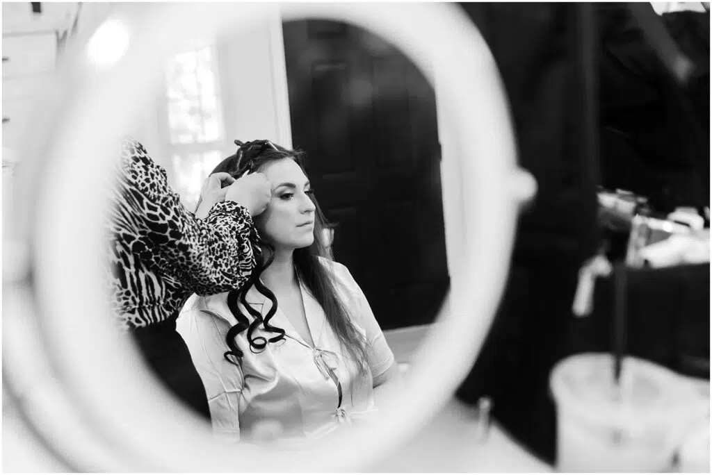 Snohomish Wedding Photographer GSquared Weddings In a black and white image of timeless elegance, a woman preparing for a summer wedding in Port Gamble sits in front of a round mirror. She wears a robe, her hair partially curled by another, while styling tools and a cup clutter the counter. Serving Seattle, Snohomish and Orlando