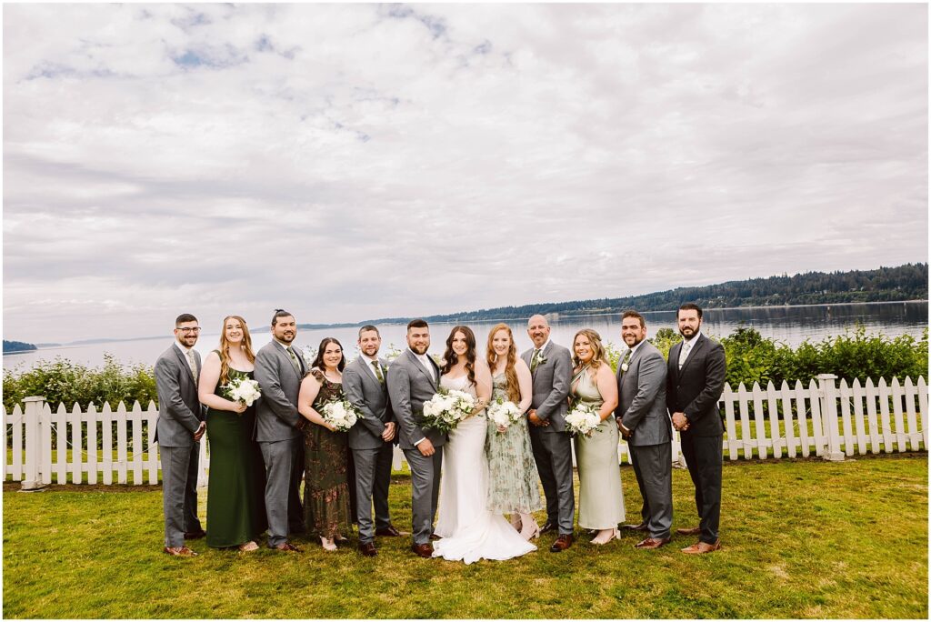 Snohomish Wedding Photographer GSquared Weddings A wedding party poses in front of a white picket fence in Port Gamble, with a scenic lake and forested hills as the backdrop. Dressed in formal attire and holding flowers, the group stands on the grassy area. The sky is cloudy, capturing the essence of a summer wedding. Serving Seattle, Snohomish and Orlando