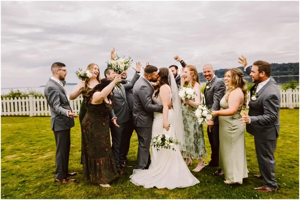 Snohomish Wedding Photographer GSquared Weddings A bride and groom share a kiss in the center of a joyful summer wedding party. The group, dressed in suits and dresses, celebrates with raised arms and bright smiles. A white fence and the overcast sky of Port Gamble serve as the backdrop. Serving Seattle, Snohomish and Orlando