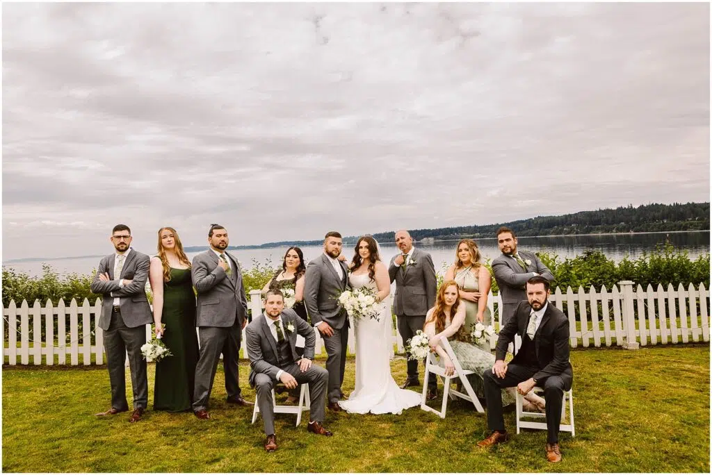 Snohomish Wedding Photographer GSquared Weddings A wedding party poses outdoors in front of a white fence, capturing the charm of a Port Gamble summer wedding. With scenic views of water and trees, the group features seven men in gray suits and five women with bouquets, while the bride and groom stand proudly in the center. Serving Seattle, Snohomish and Orlando